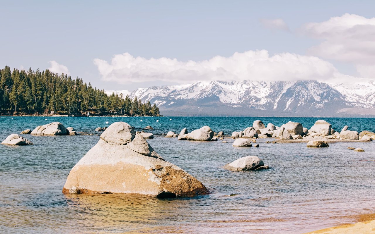 Granite boulders along Lake Tahoe, with forested shores and snow-covered mountains beyond.