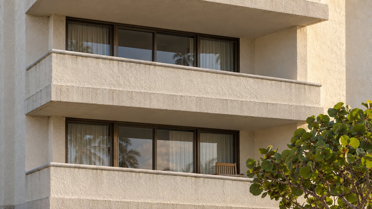 Two recessed balconies in a cream stucco condominium, with bronze sliding doors, weathered concrete edges, and sea grape leaves below.