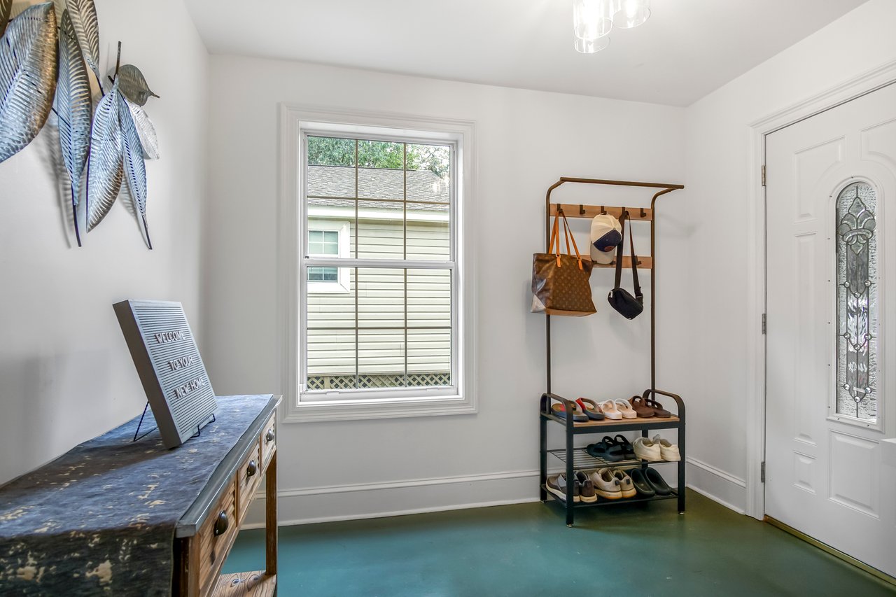 A bright, minimalist entryway with a window, a white door, a black shoe rack, and a rustic bench. Decor includes hanging plants and a wall art piece.