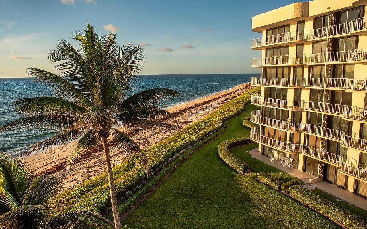 Beachfront condominium with curved balconies, landscaped grounds, palm trees, and ocean views.