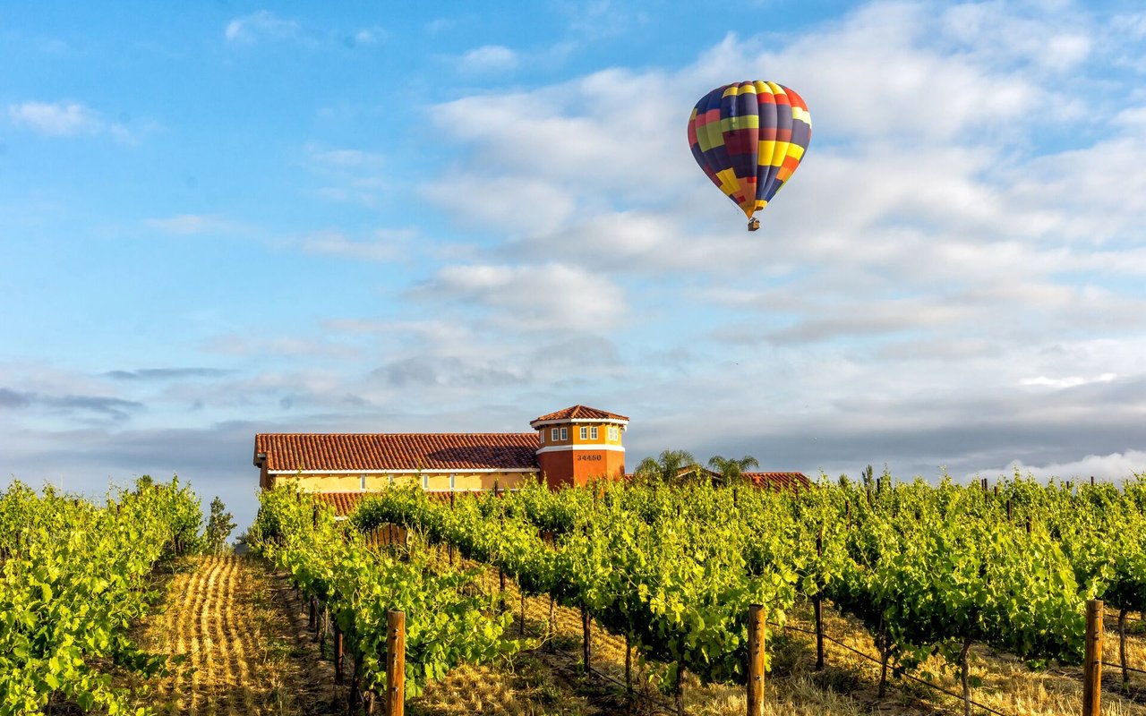 Temecula vineyard rows and a terracotta-roofed winery beneath a colorful hot air balloon.