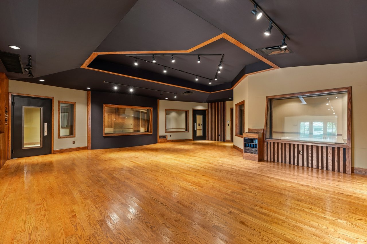 Vacant recording studio with a polished wood floor, large studio windows, and an angular ceiling.
