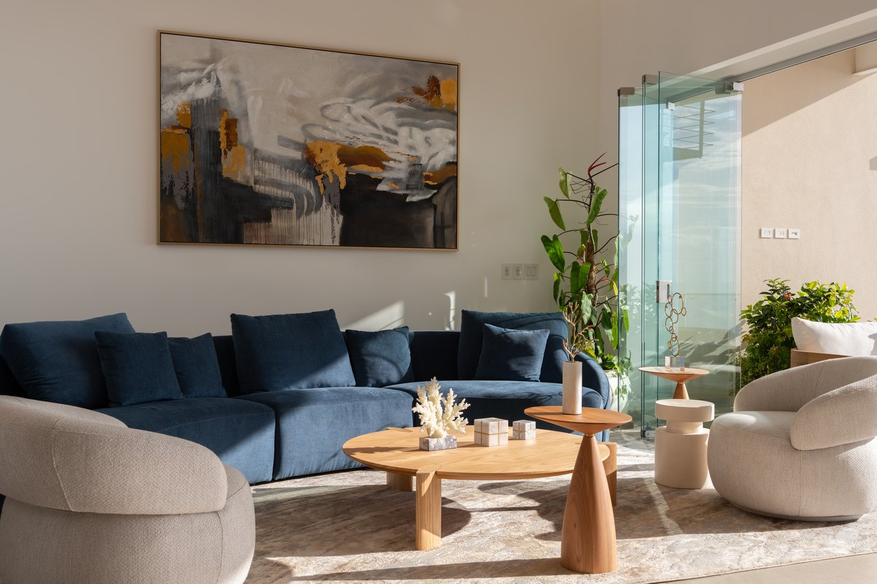 Contemporary condo living room with a blue sectional, cream armchairs, wooden tables, and glass balcony doors.