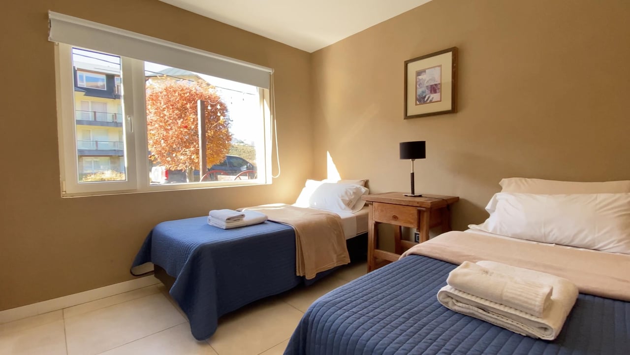 Bedroom with two single beds, blue quilted bedspreads, folded towels, and a sunlit window.