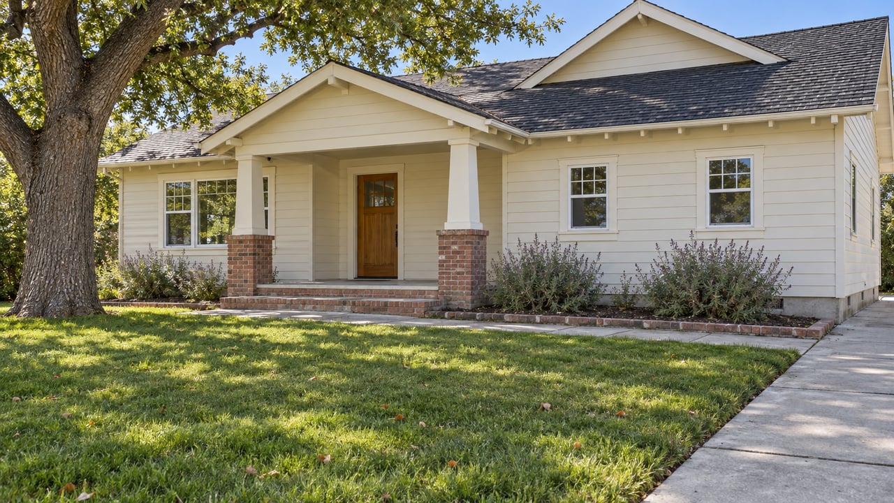 Ivory-sided single-story home with a brick-step porch, deep green front lawn and mature oak in bright daylight.