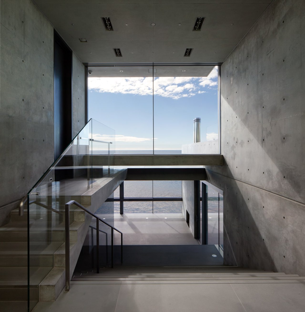 Modern concrete staircase with glass railings and ocean views through floor-to-ceiling windows.