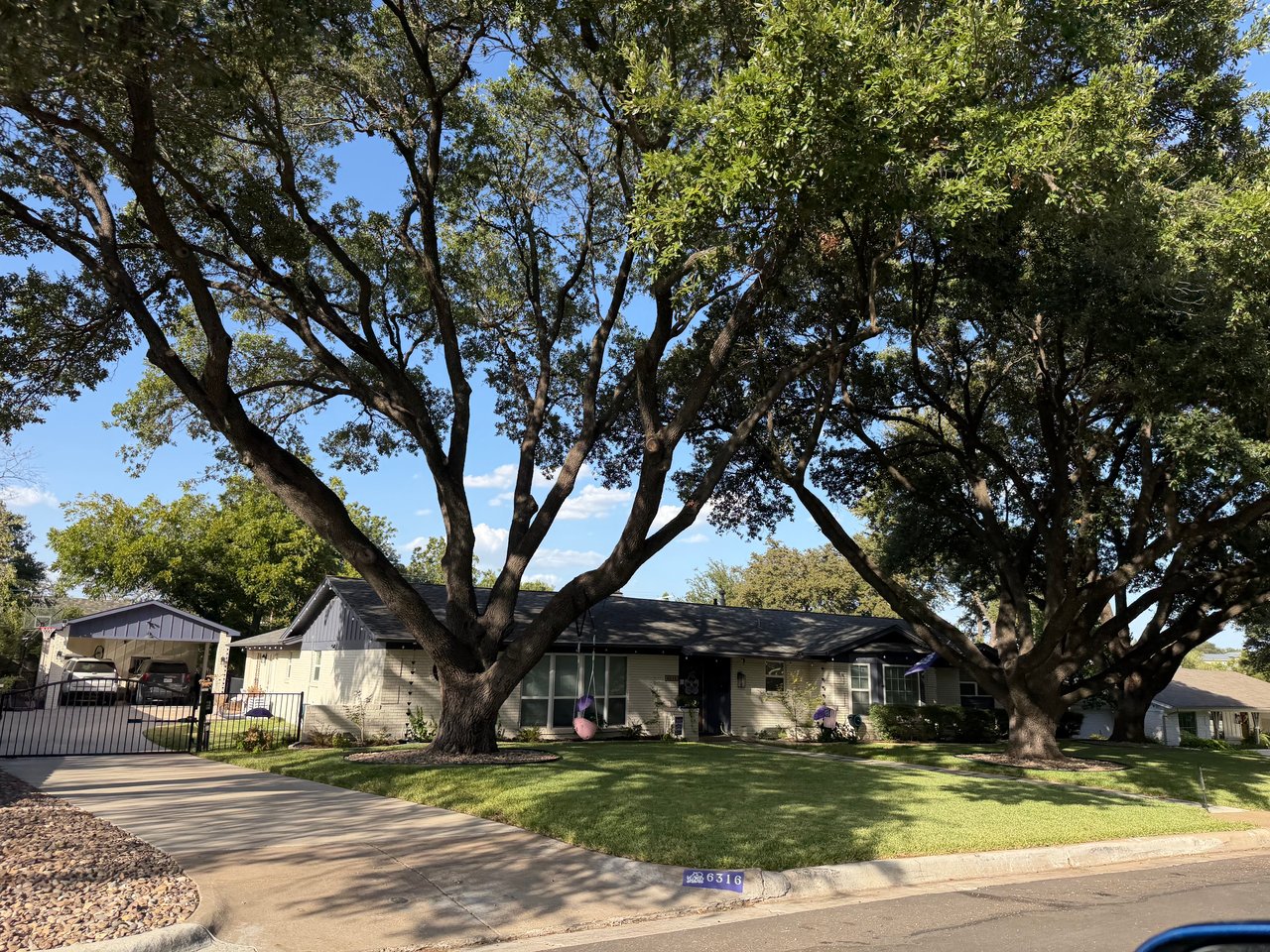 Single-story home shaded by two large trees, with a gated driveway and broad front lawn.