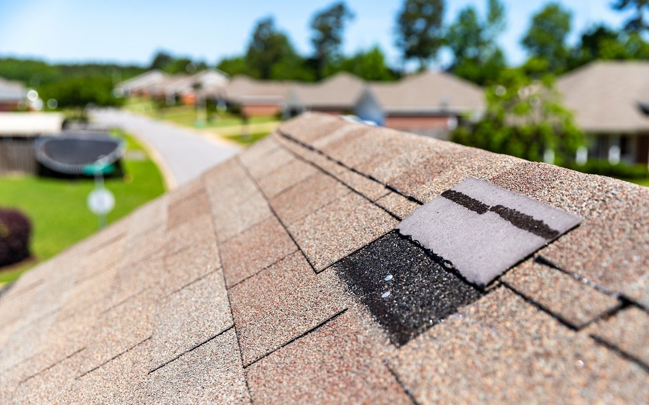 Damaged asphalt shingles expose a dark section of roof above a suburban neighborhood.