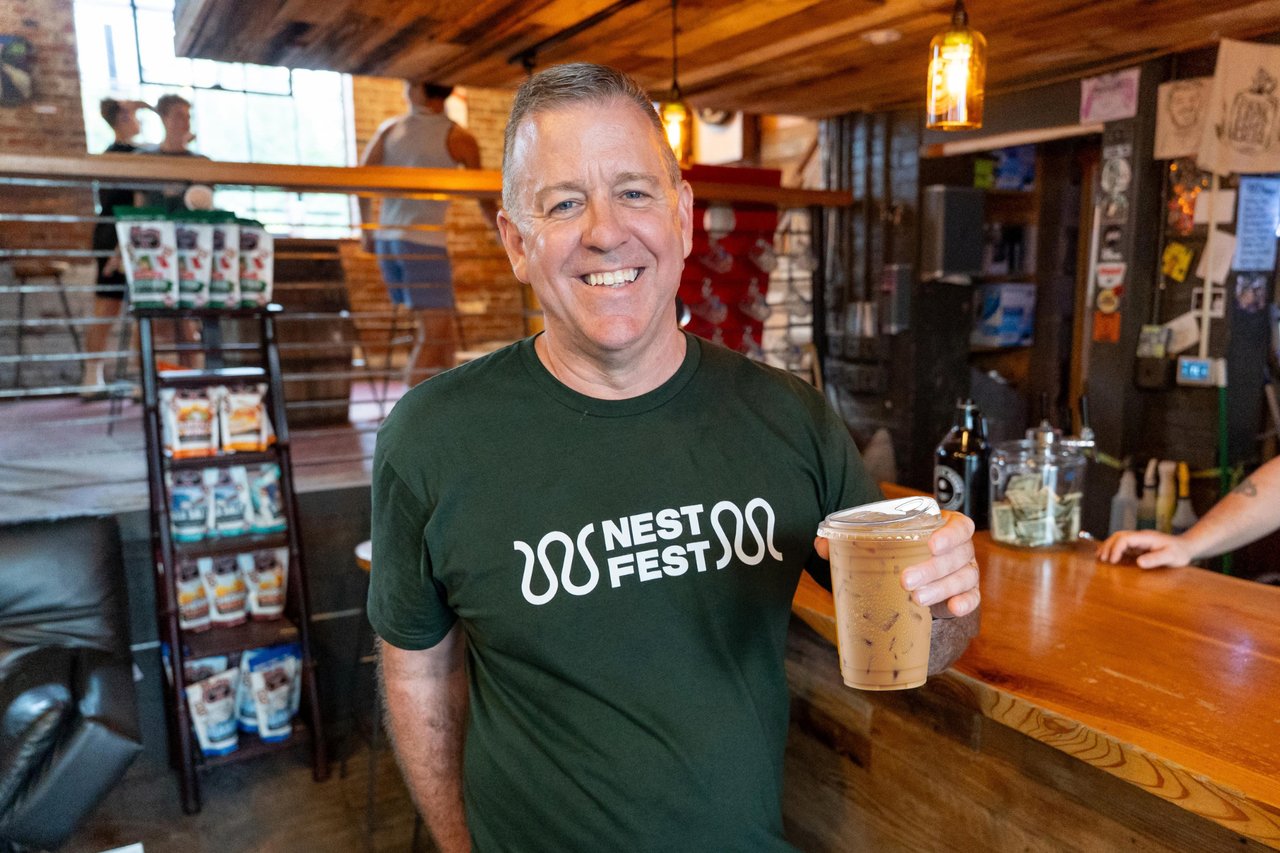 Smiling man in a green NEST FEST shirt holding an iced drink at a café counter.