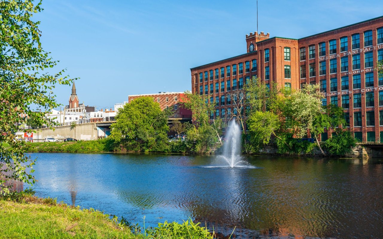 Downtown Nashua mill building and church steeple beside a river with a fountain.
