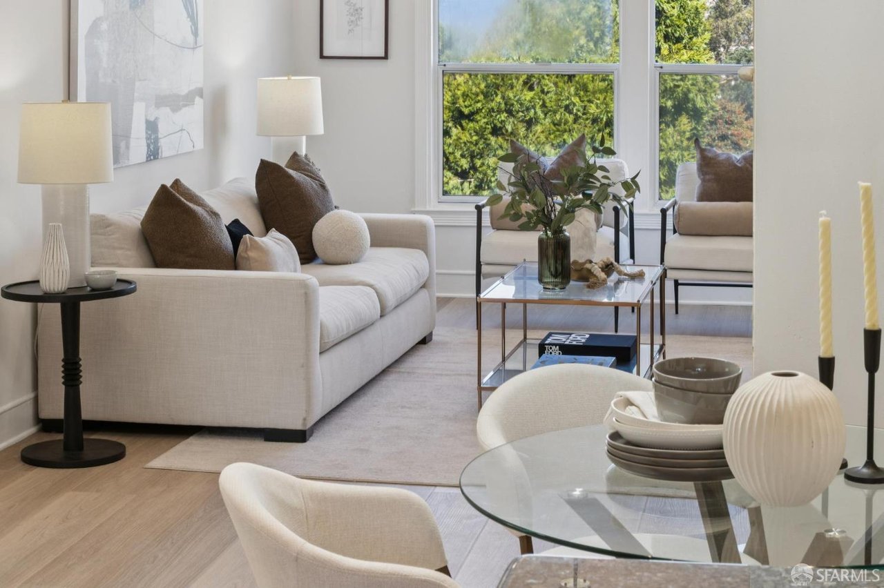 Staged living room with a cream sofa, glass coffee table, and tall windows overlooking greenery.