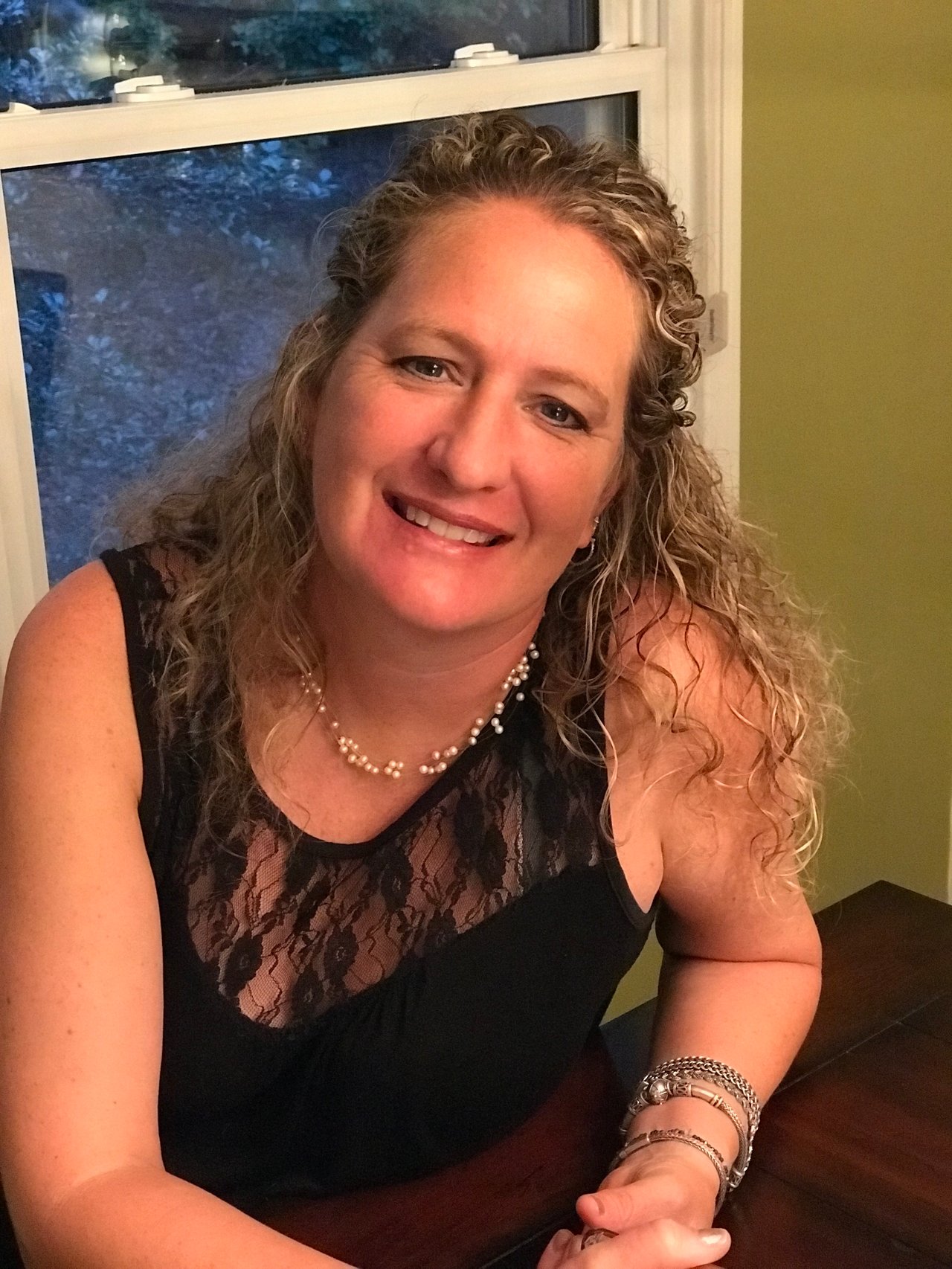 Melissa Boney smiling beside a window, wearing a black lace-trimmed top and pearl necklace.