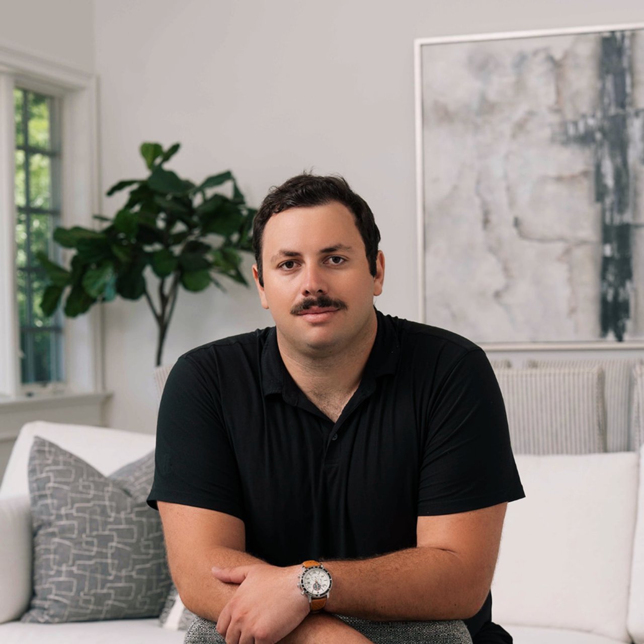 Nathan Niculae seated on a white sofa in a black polo shirt, with a leafy plant behind him.
