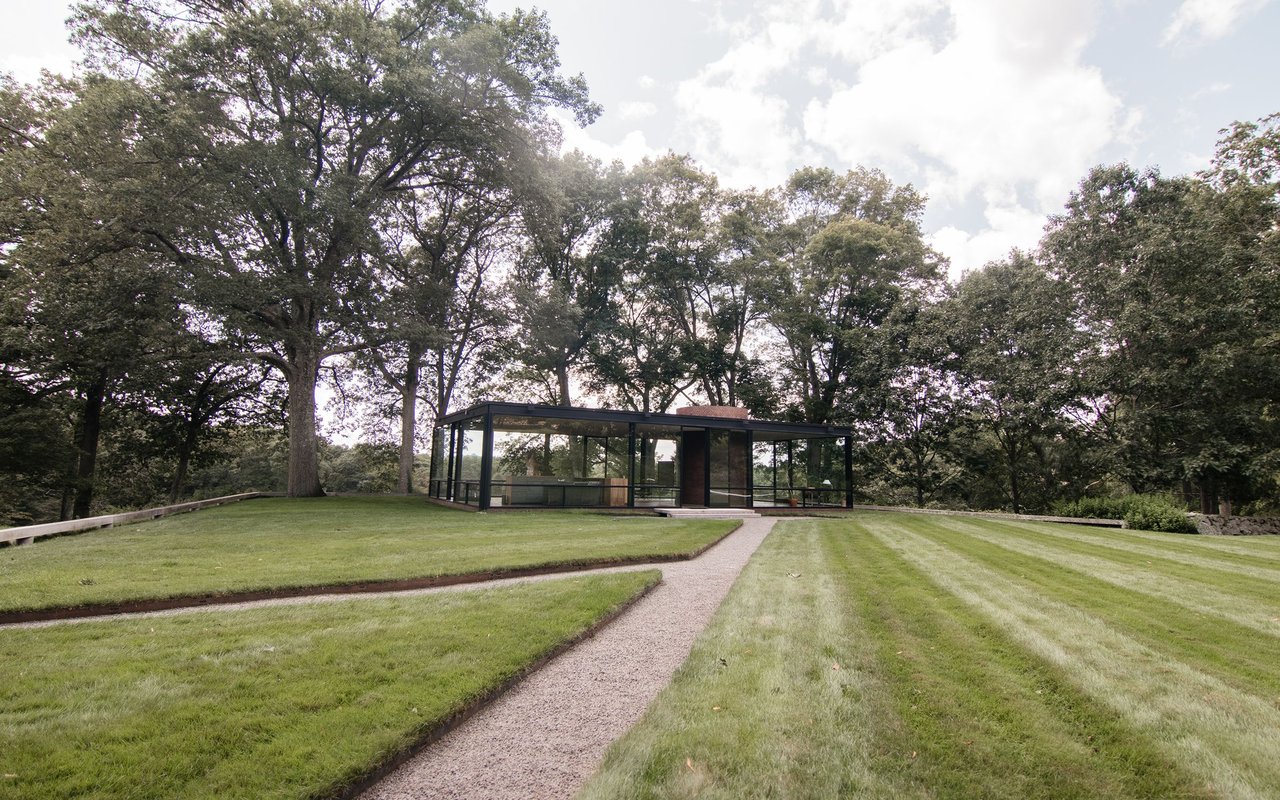 Modern glass-walled home on a broad lawn, approached by a gravel path beneath mature trees.