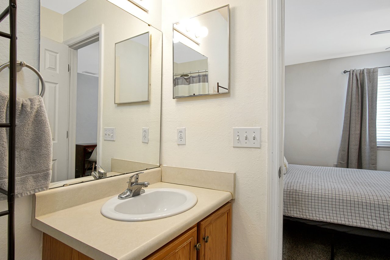 A bathroom with a countertop sink, mirror, and beige walls. A towel hangs on a rack. The adjacent bedroom features neutral decor and curtains.