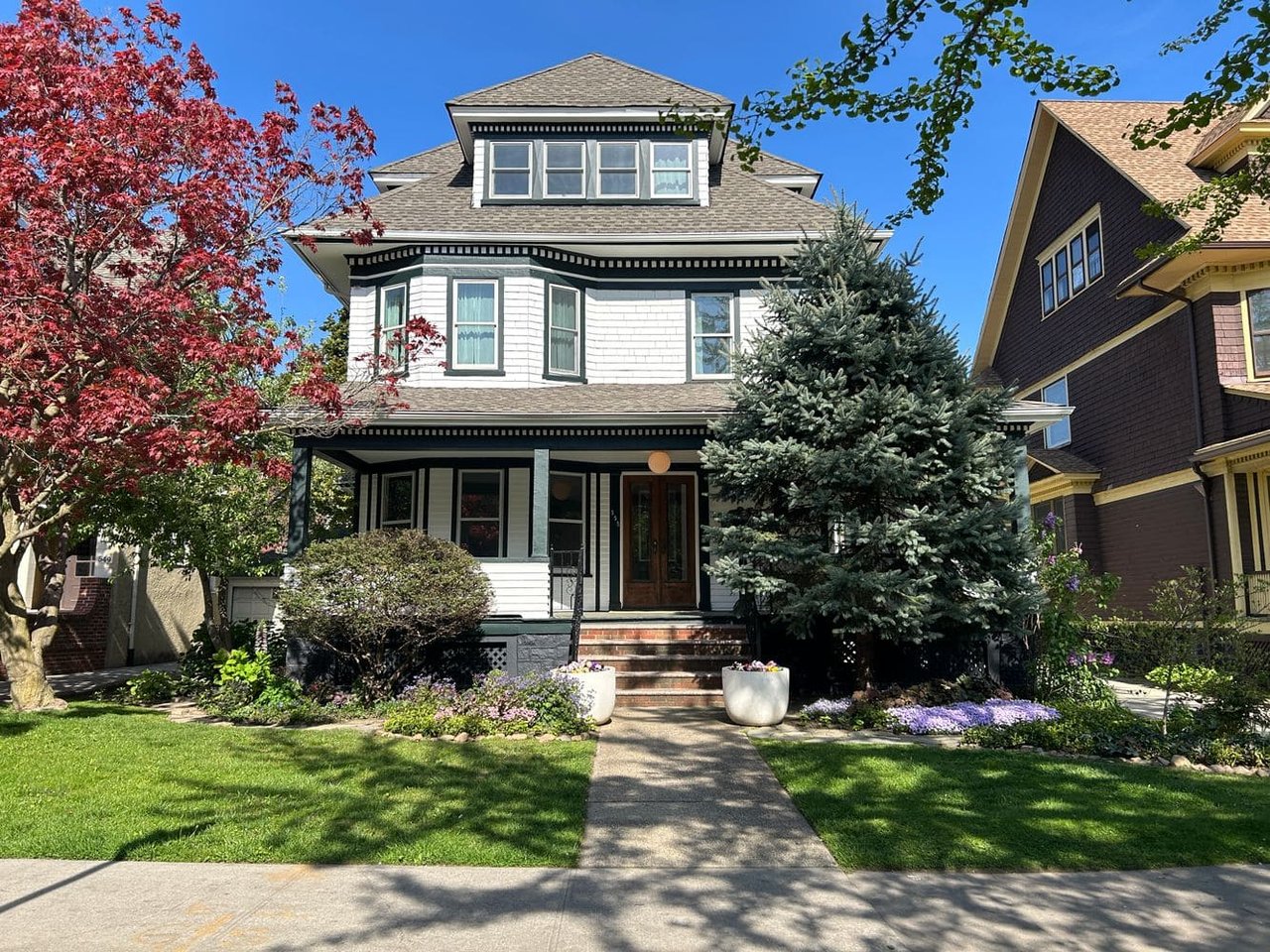 Three-story white house with gray roof, front porch, surrounded by trees and a well-maintained garden, with a sidewalk leading to the entrance