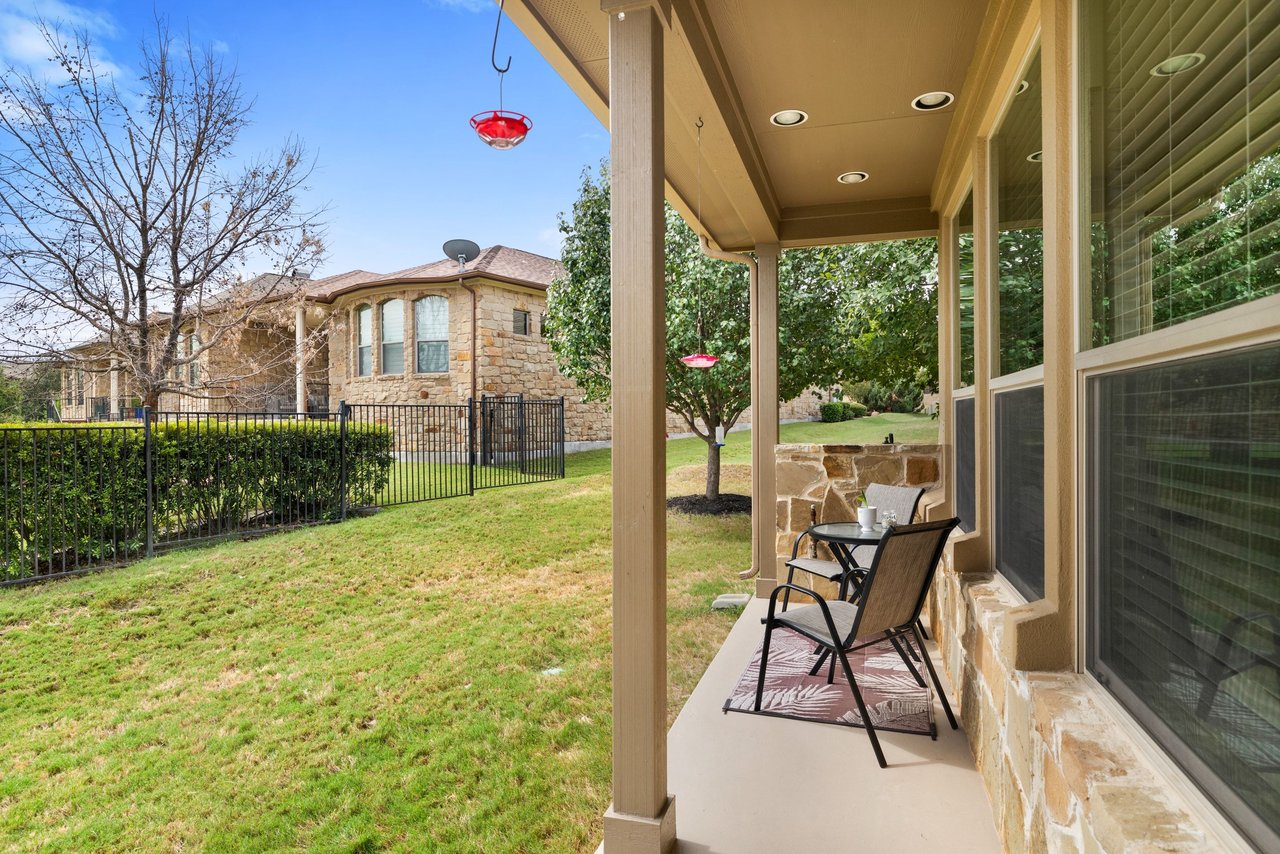 Covered patio with two chairs overlooks a fenced grassy yard beside a stone home.