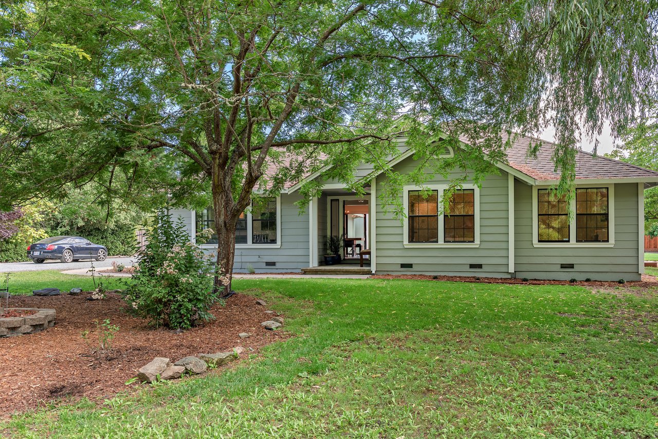 Sage-green single-story house framed by mature trees, with a covered entrance and broad front lawn.