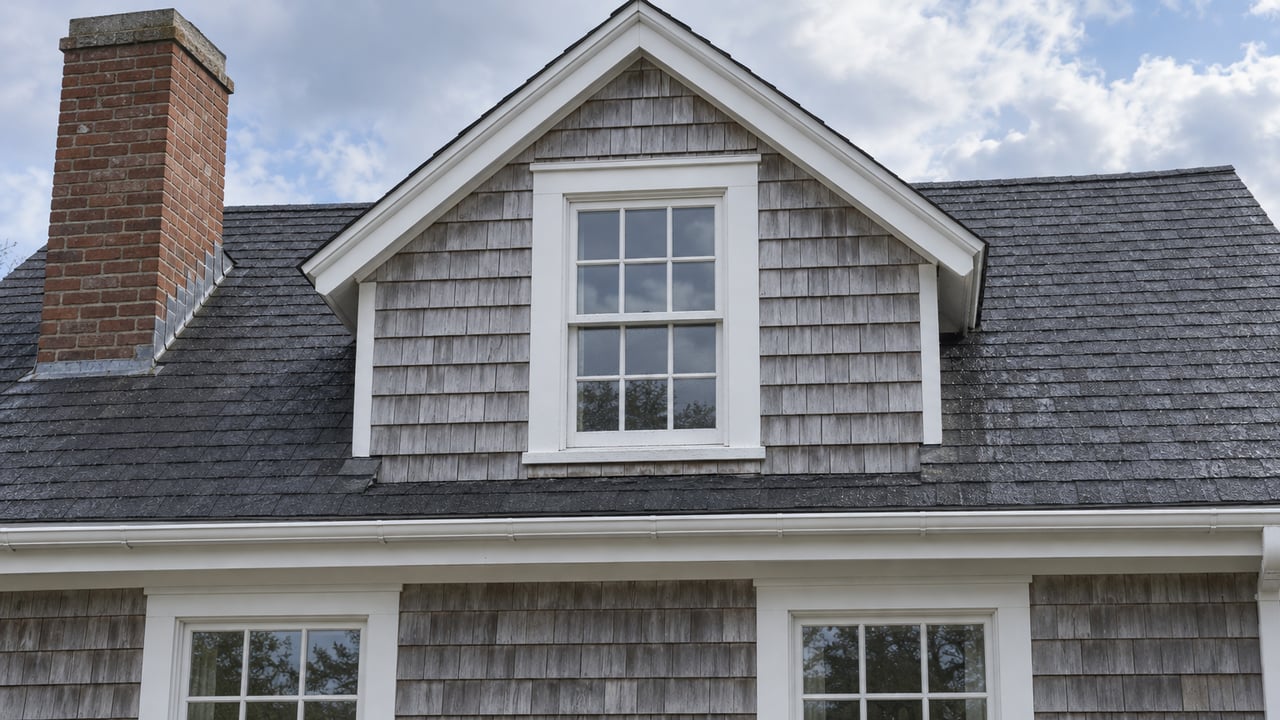 Weathered cedar shingles, a white-trimmed dormer window, and a brick chimney beneath soft post-rain daylight.
