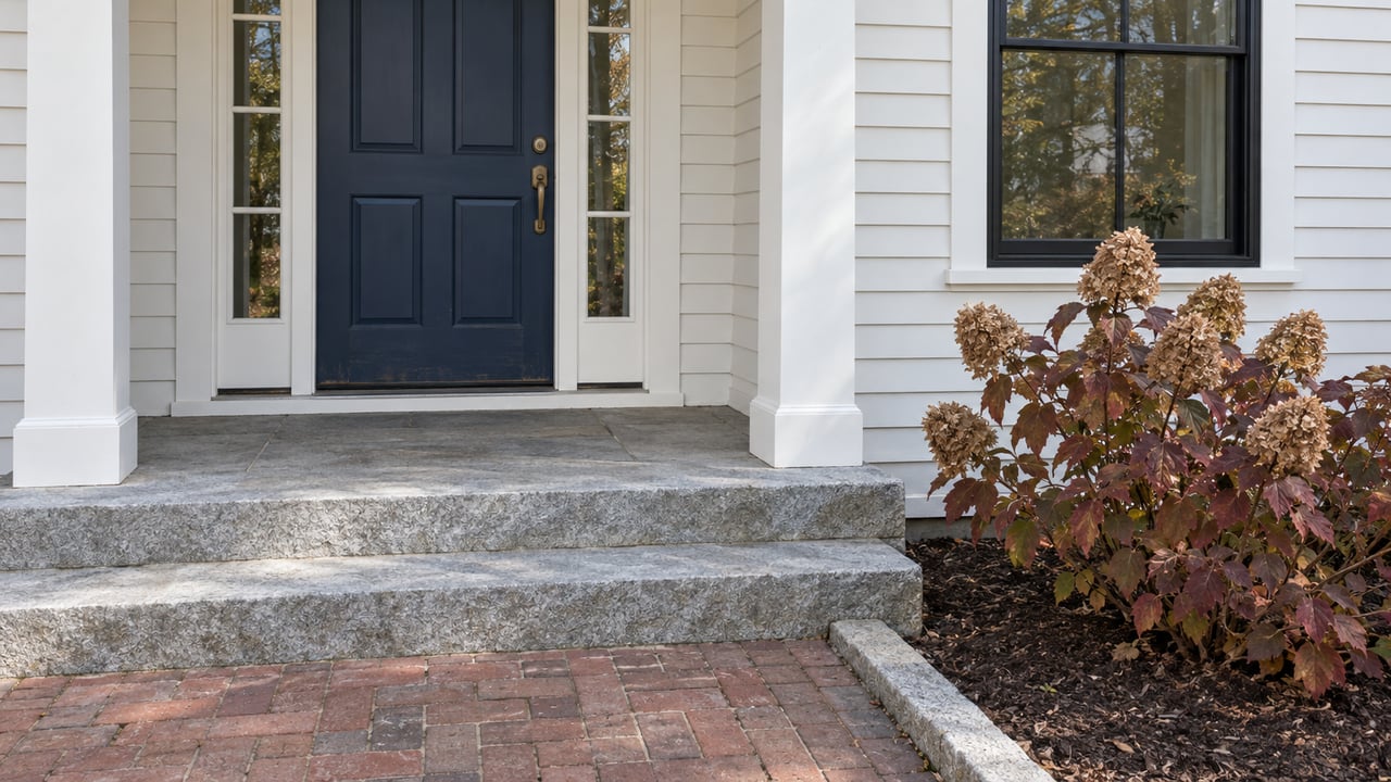Autumn light on a white clapboard home's navy entry door, gray granite steps, brick walk, and burgundy oakleaf hydrangea.