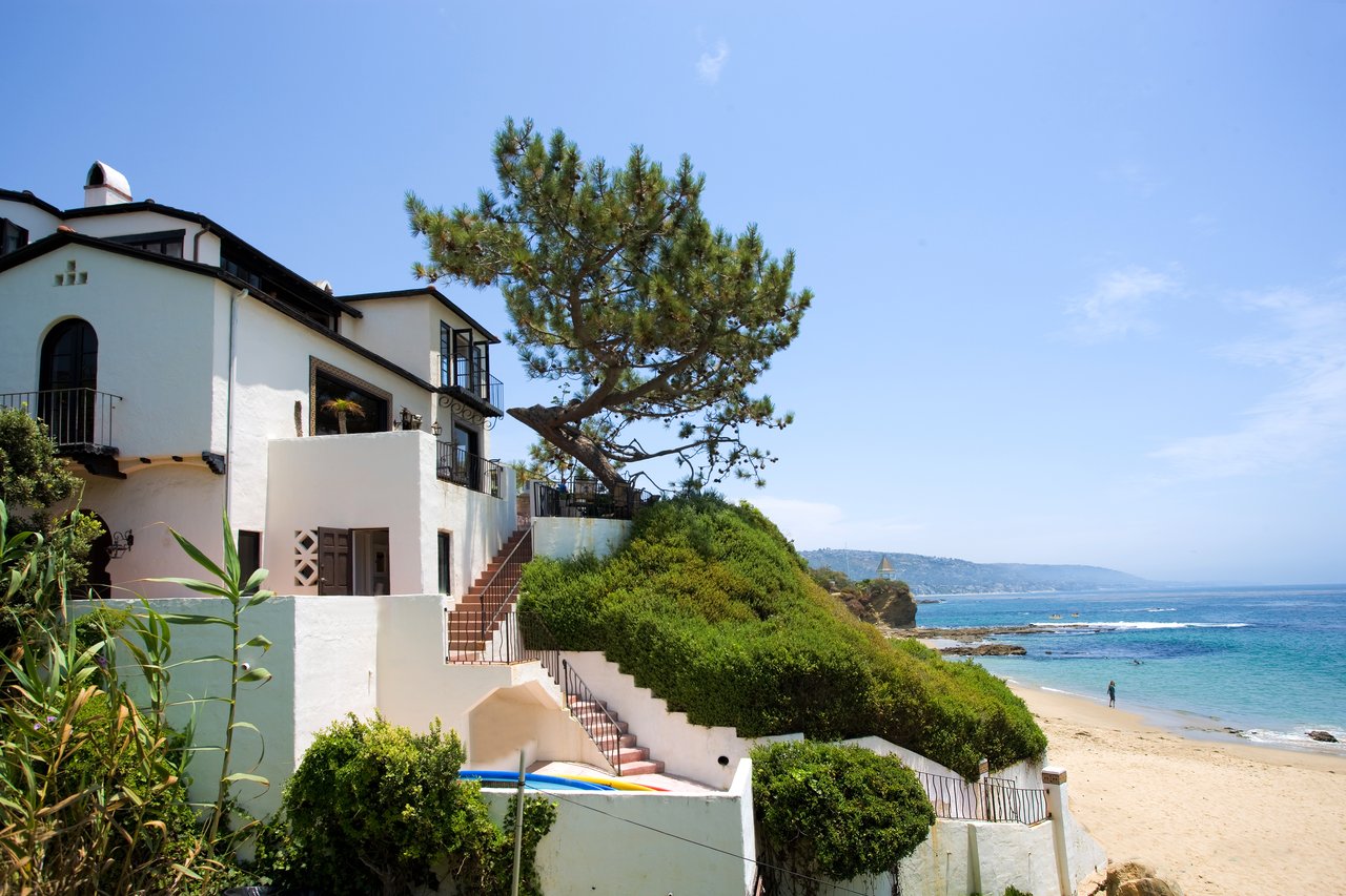 White coastal home above a sandy beach, with ocean views and stairways through the greenery.