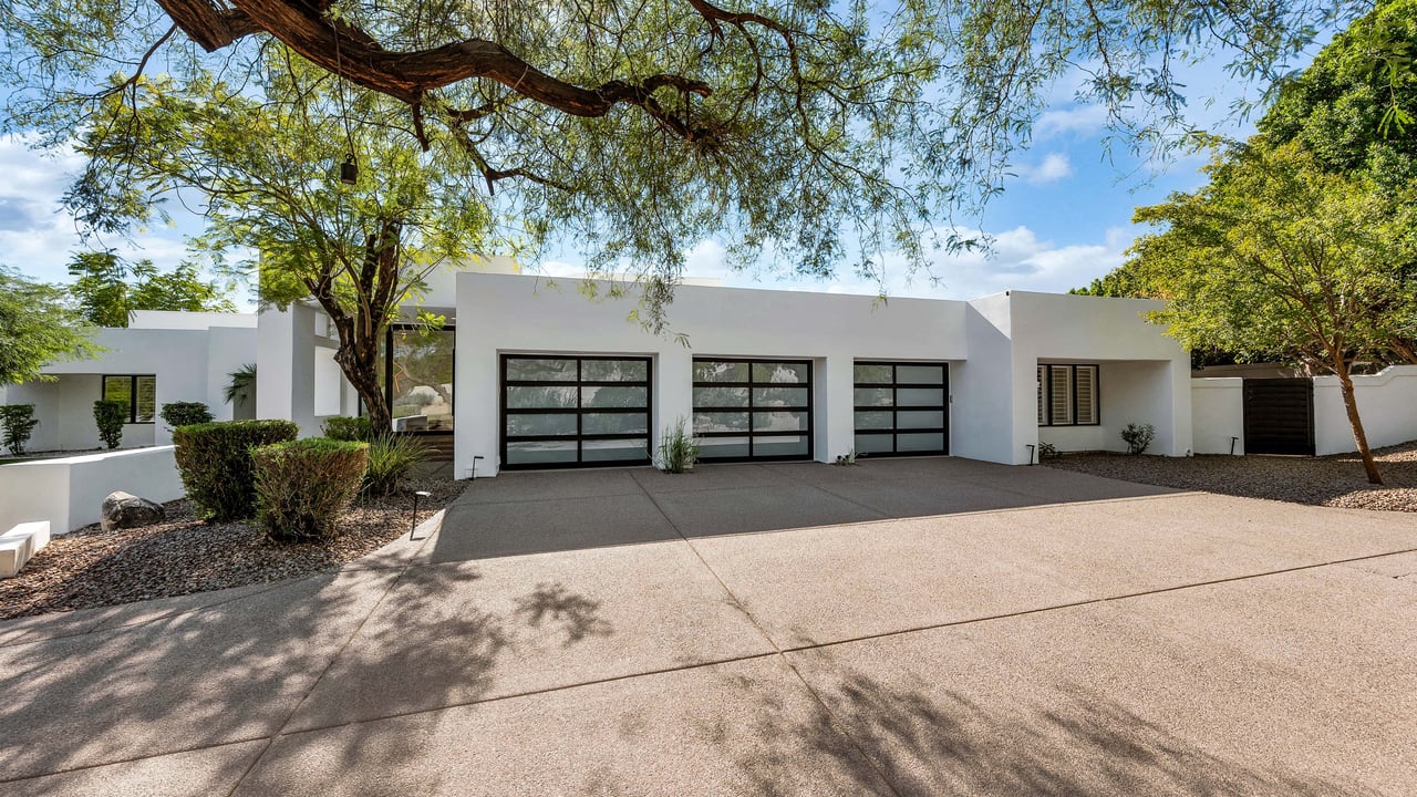 Modern white home with three black-framed garage doors, a wide driveway, and mature shade trees.