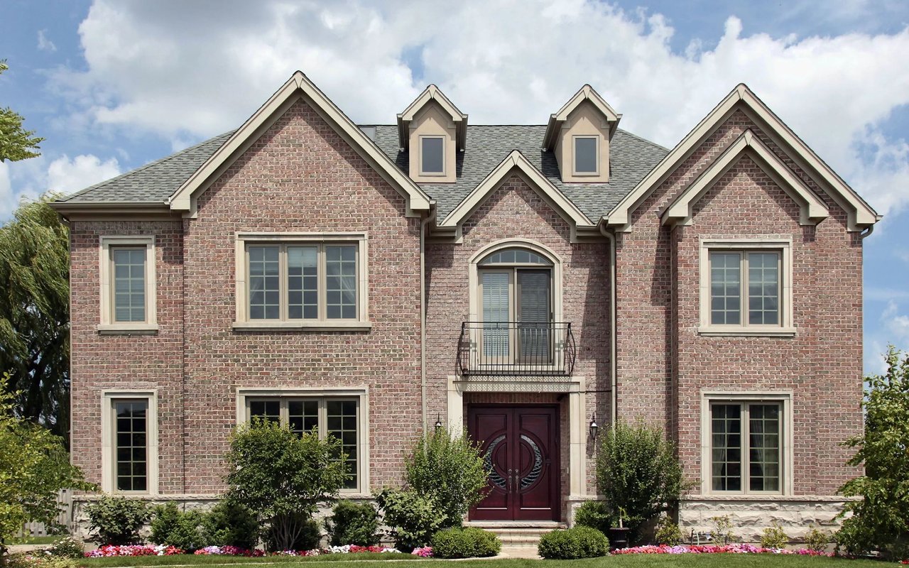 Red brick house with multiple gables, a balcony, dark red double doors, and landscaped flower beds.
