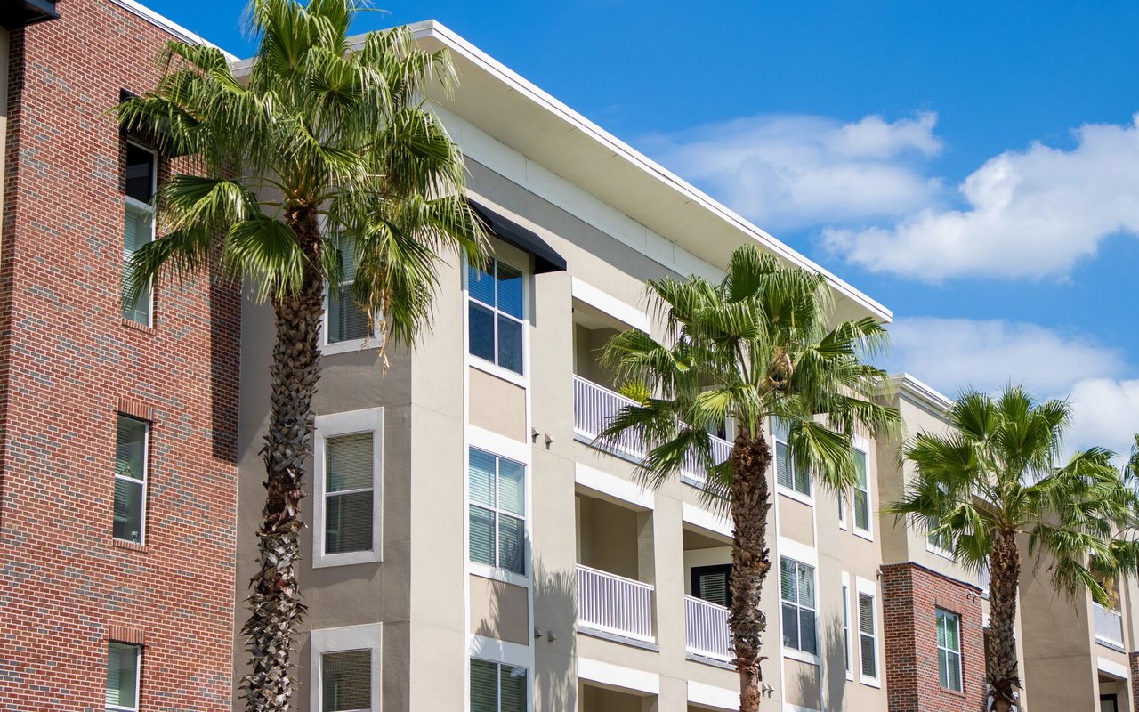 Multi-story residential building with white balconies, red brick sections, and palm trees.