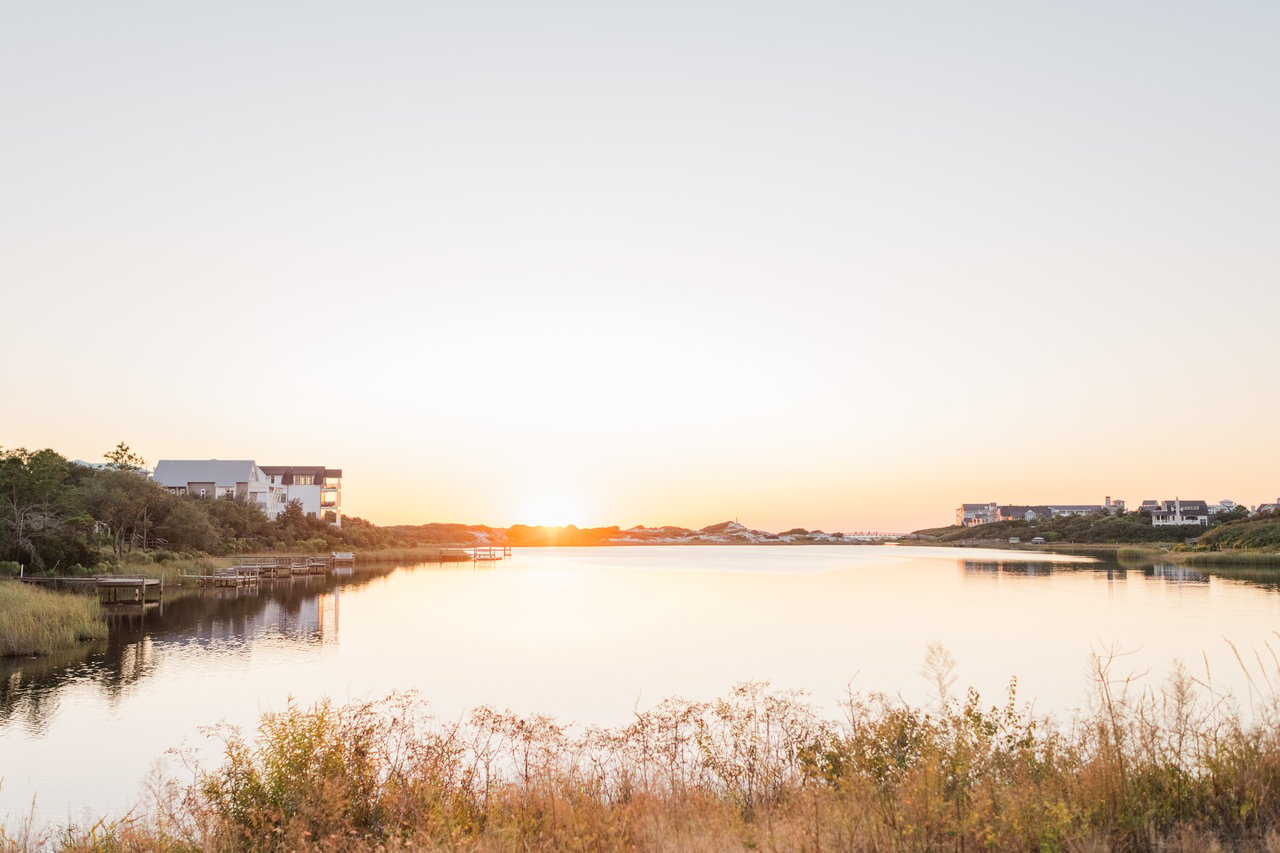 A stunning view of the Seagrove dune lake on 30A, surrounded by luxury homes, with the sun setting over the sand dunes in the background, casting a golden glow on the water.