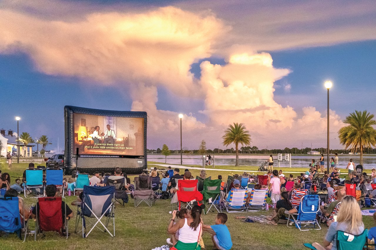 Families watch an outdoor movie beside a lake at dusk, seated in lawn chairs.