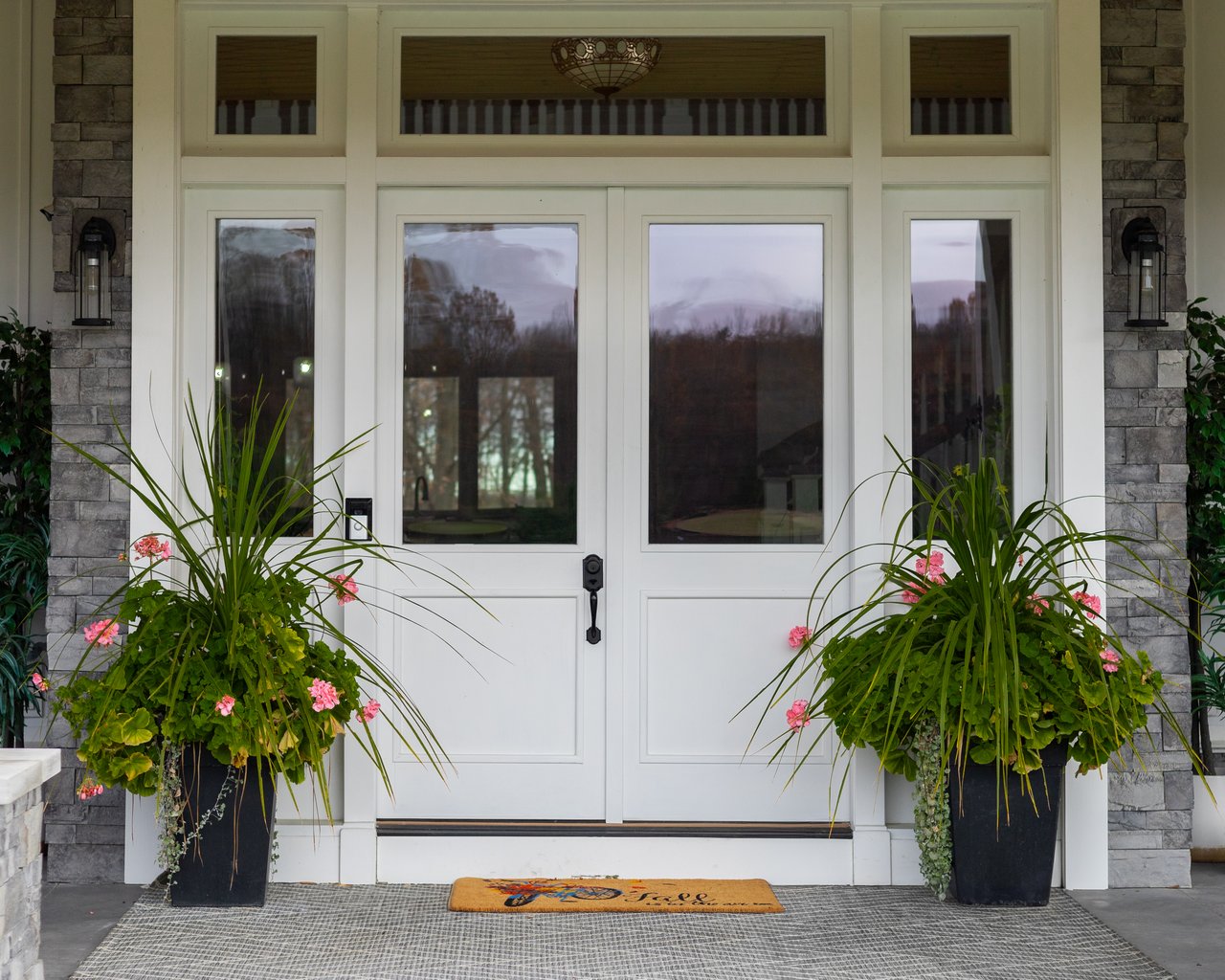 White double front doors framed by stone, with flowering planters and an orange doormat.