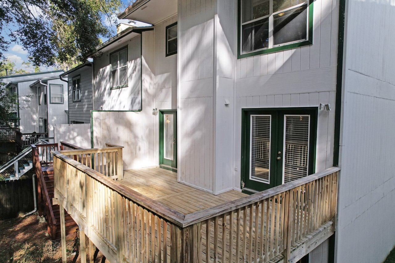 Rear view of the townhome at 529 Collinsford Road, Tallahassee, Florida, showcasing a spacious elevated wooden deck with railings and dual French doors framed in green trim. The deck provides access to the main living area and offers a private outdoor space shaded by mature trees.