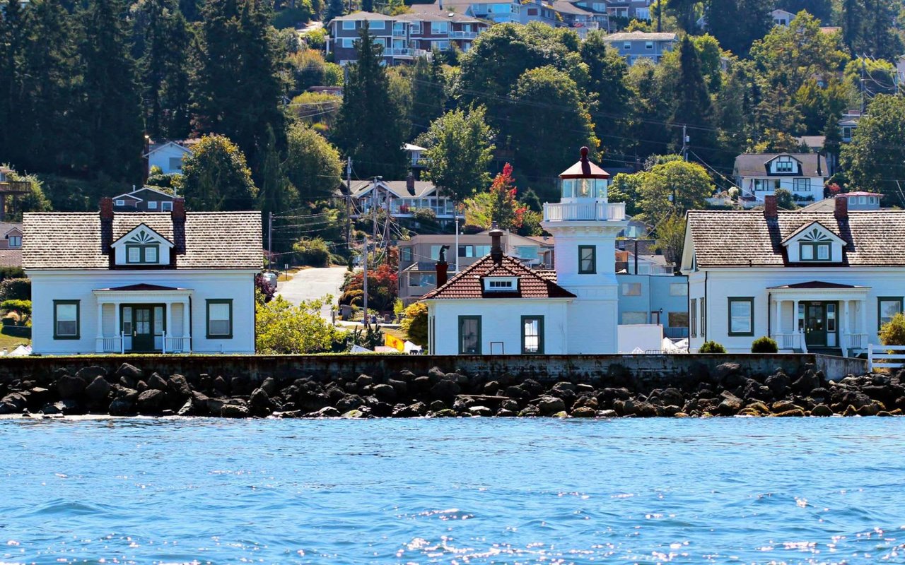 White waterfront buildings with a cupola stand above a rocky seawall, with wooded hills behind them.