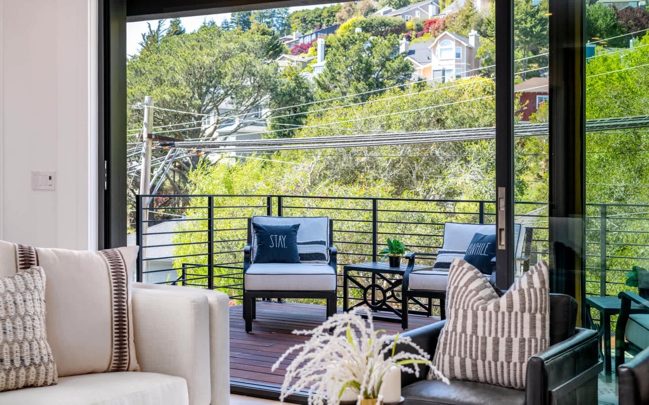 Furnished living room opens onto a balcony with lounge chairs and a leafy hillside view.