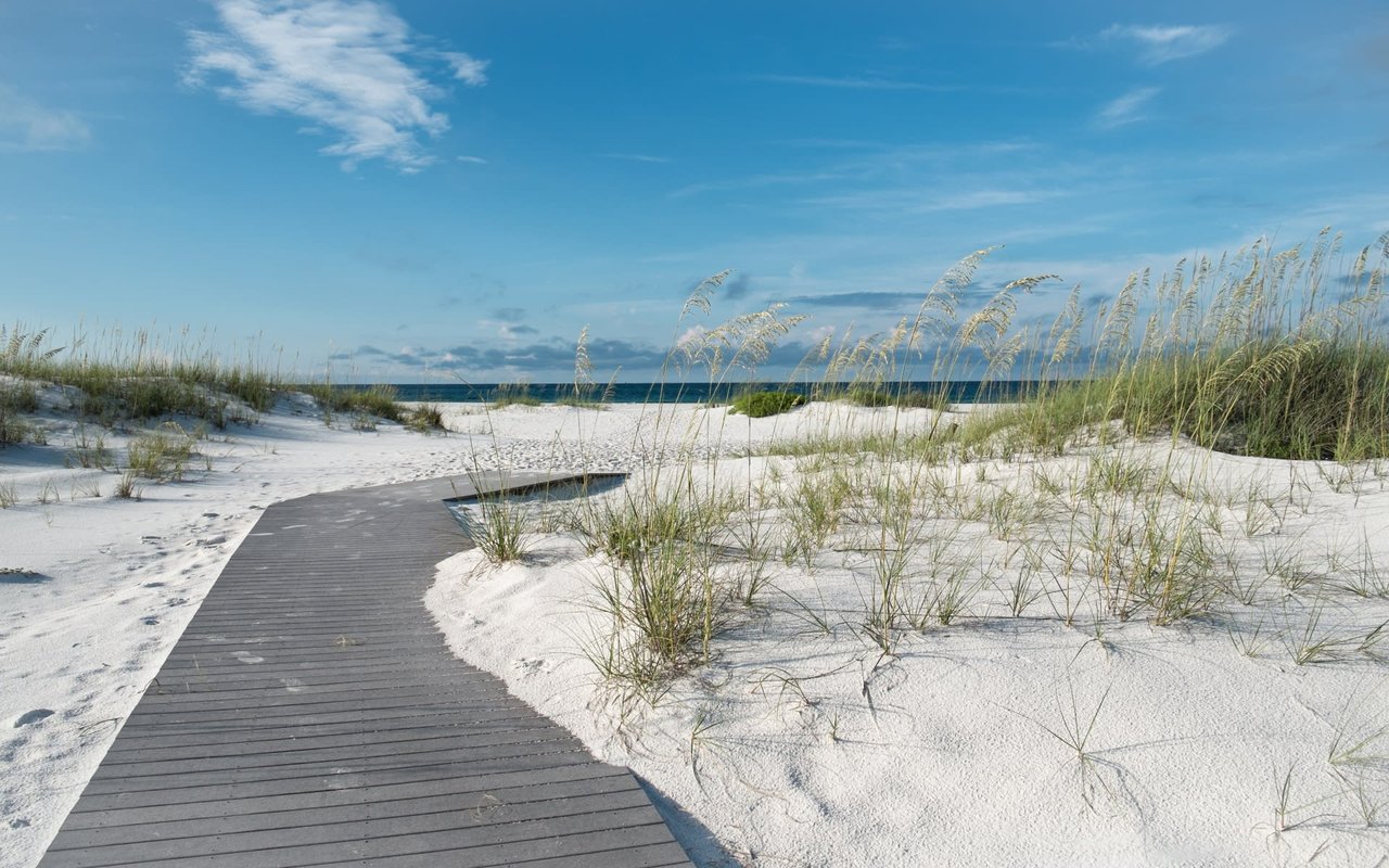 A wooden boardwalk winds through powdery white sand dunes toward a pristine turquoise beach under a vibrant blue sky.