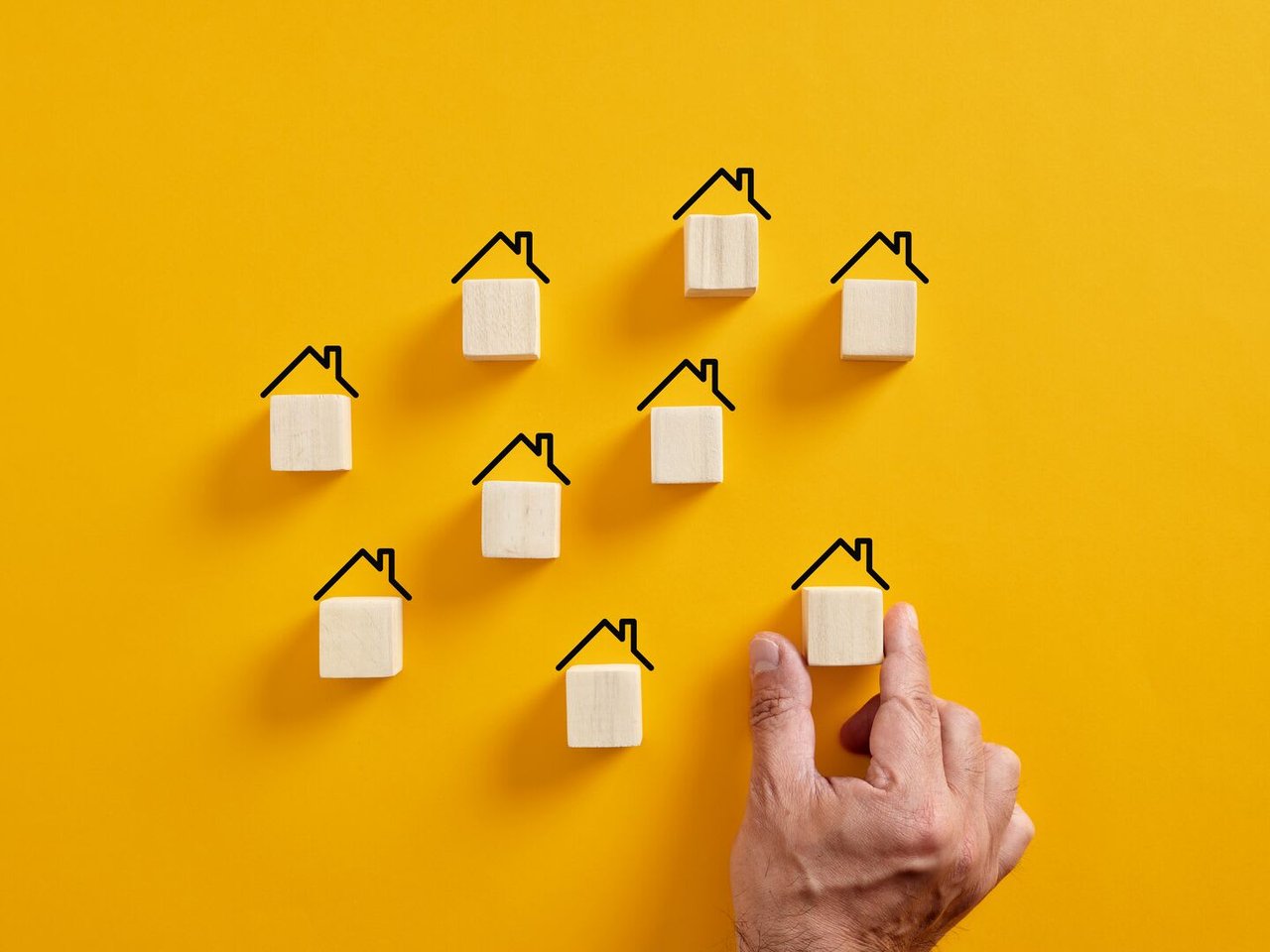 A yellow background featuring miniature house models and a person’s hand arranging them. The scene suggests a concept related to real estate or housing.