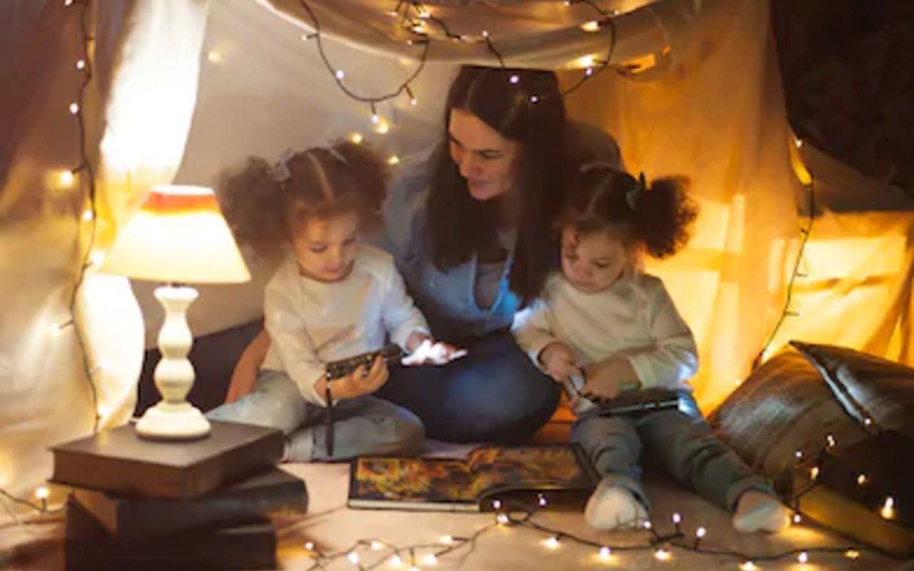 Woman and two children relax inside a blanket fort lit by string lights.