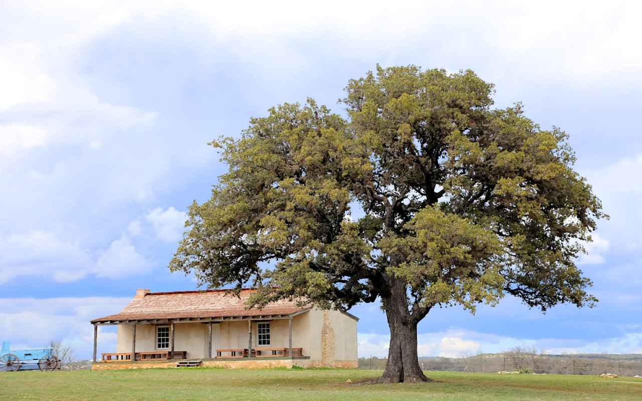  A historic-looking, single-story house with light beige walls and a reddish-brown tile roof, situated in a grassy field 