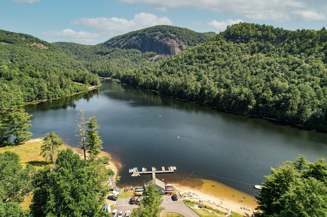 Alt=A serene lake surrounded by lush green forest and mountains under a partly cloudy sky. At the lake's shore, a small beach area is visible along with a dock and several parked boats. People, possibly real estate agents taking a break, are seen enjoying the water and the beach.