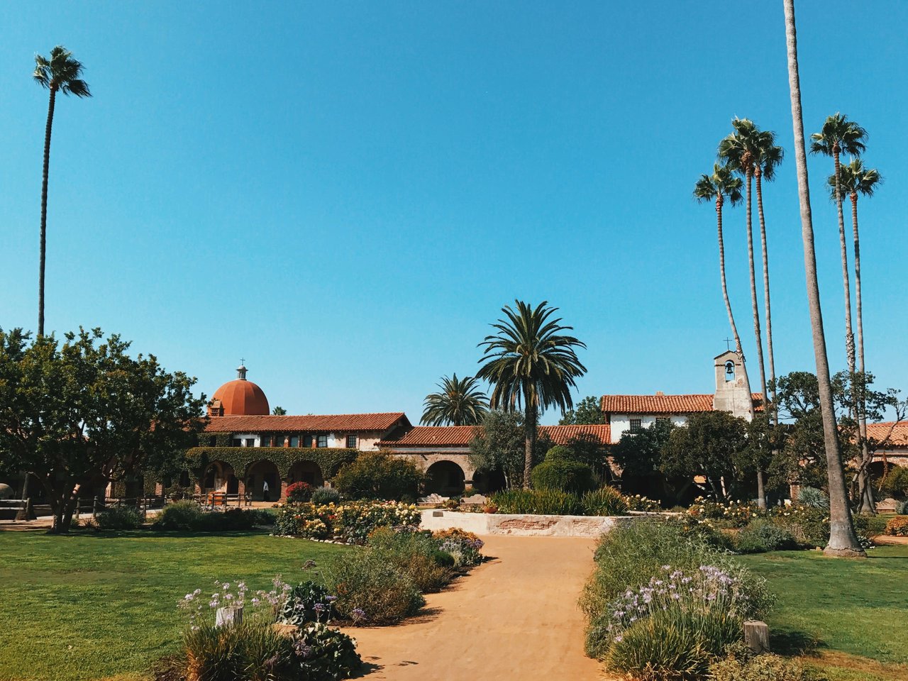 San Juan Capistrano Mission church with patches of green grass and flowerbeds along the walkway and surrounded by palm trees.