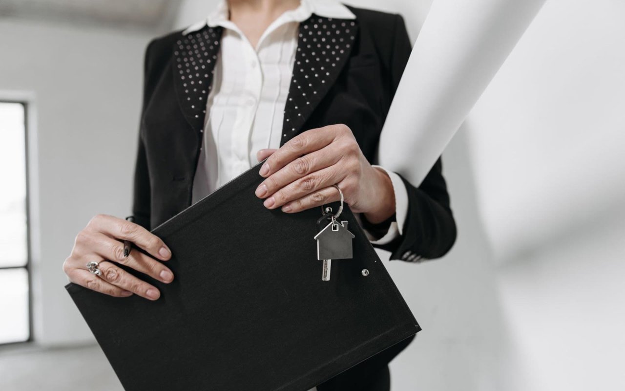 Close-up of a real estate agent holding a folder, house key, and rolled blueprint, wearing a black suit.