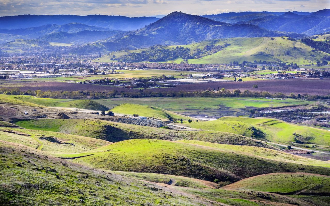 A valley with scattered houses, fields with green patches, and mountains in the background.