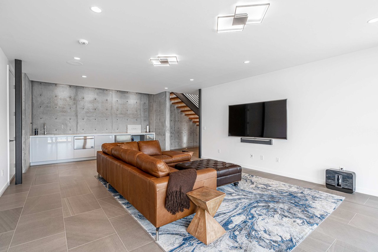 A spacious and modern living room featuring a comfortable brown leather L-shaped sofa and a matching ottoman on a large abstract blue and white rug. 