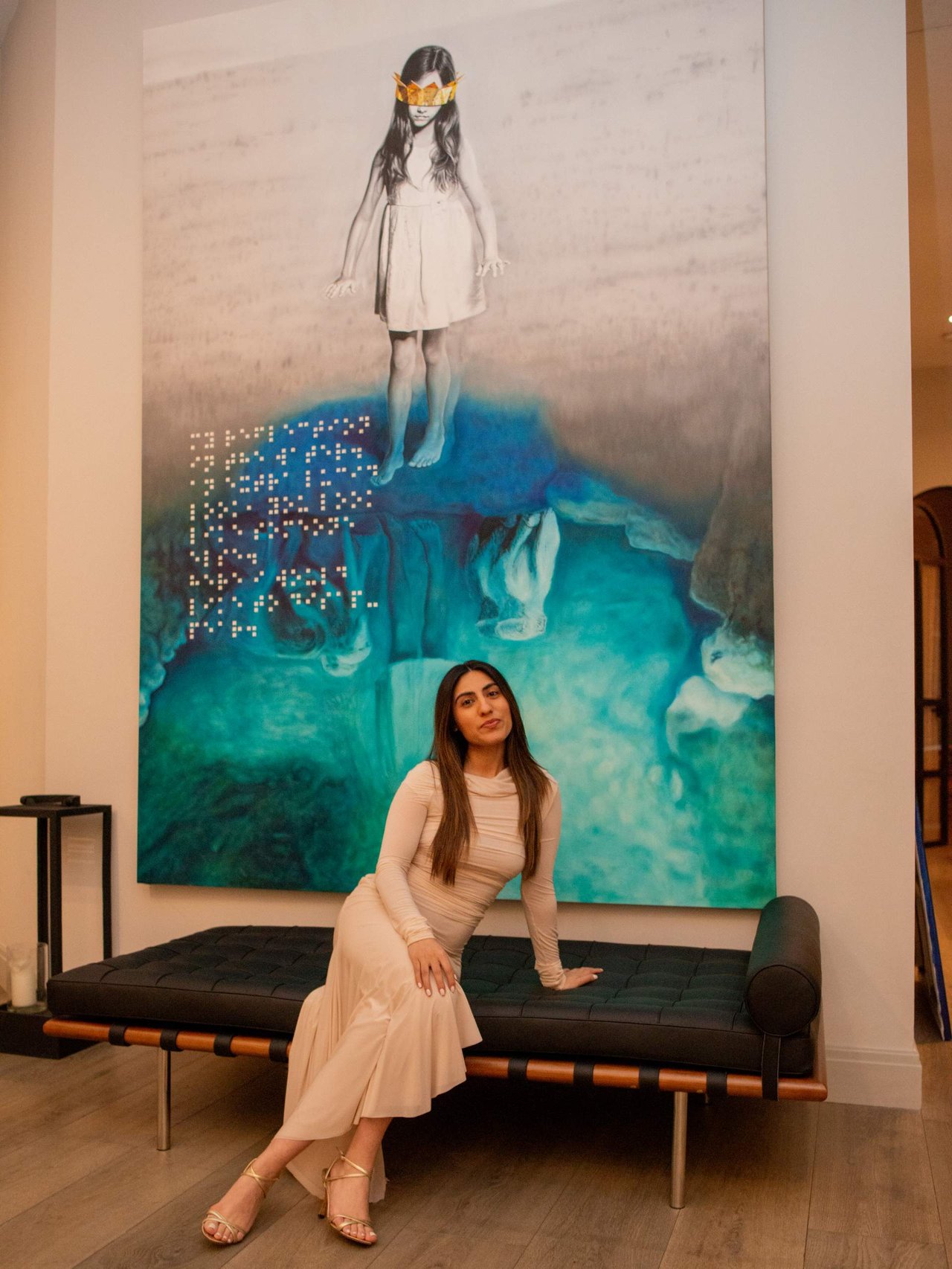 Woman in a pale dress seated on a bench beneath a large colorful artwork.