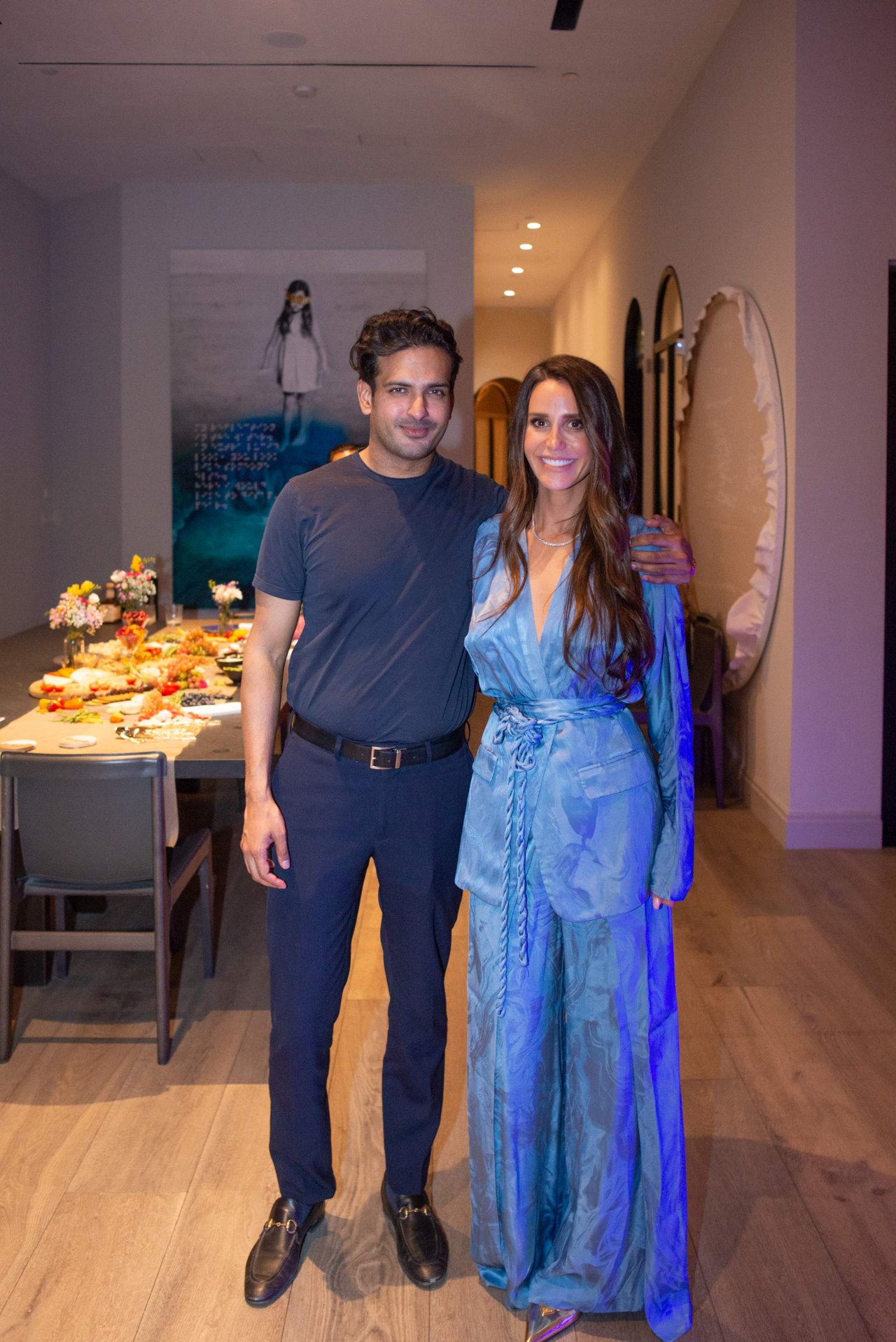 A man and woman pose beside a buffet table at an indoor event; she wears a blue patterned outfit.