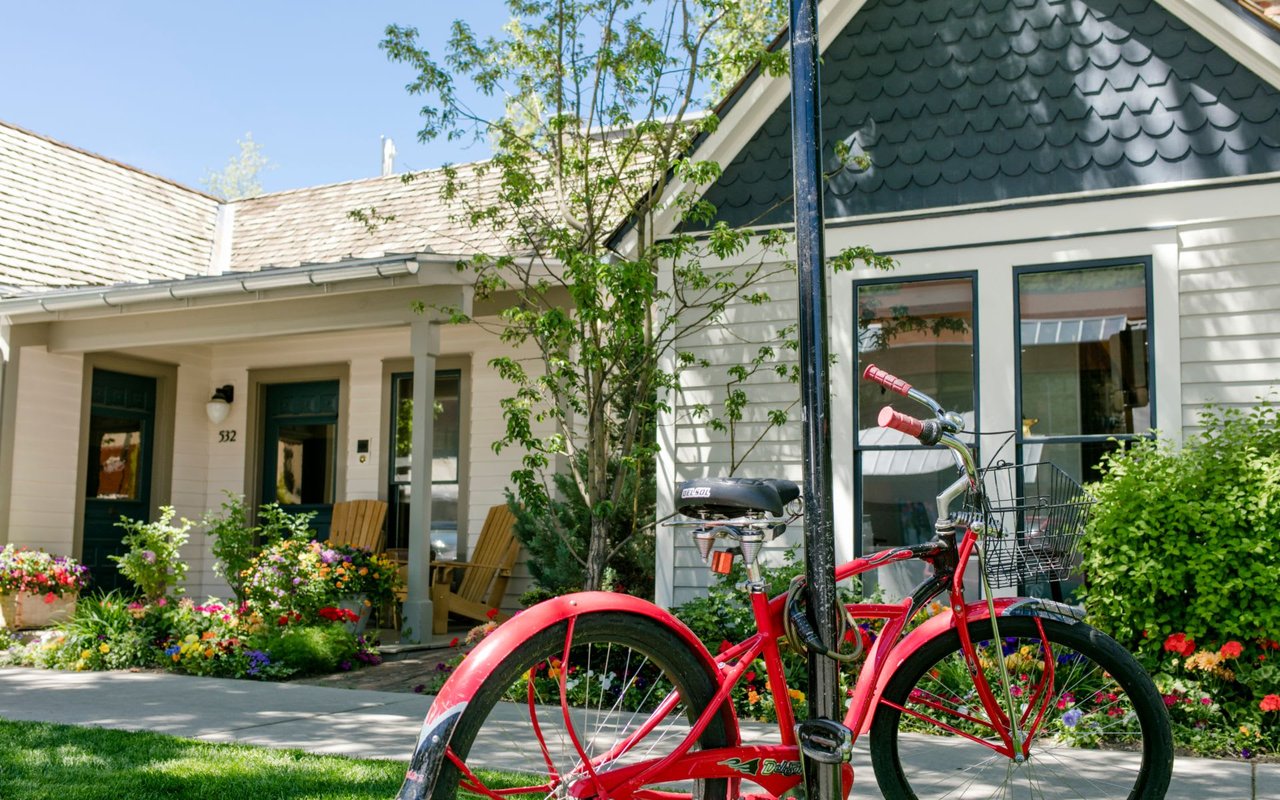 Charming white cottage in Aspen with wrap-around porch, colorful flower gardens, and a vintage red bicycle parked outside, showcasing the town's quintessential mountain lifestyle and welcoming neighborhood character