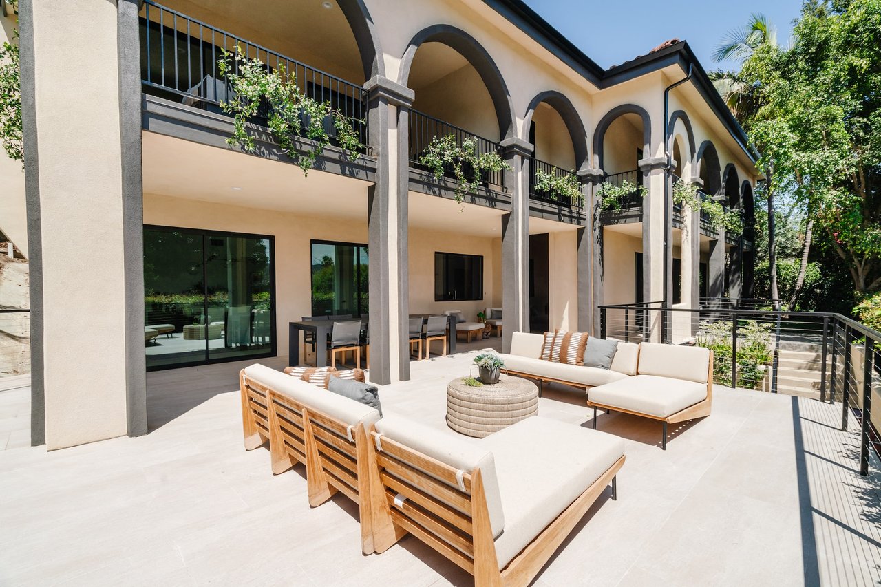  An expansive and luxurious outdoor patio extends from a large, two-story Mediterranean-style house with cream-colored walls and dark trim. 