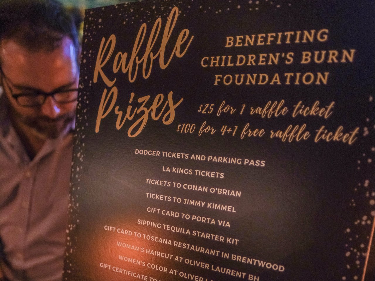  A person with glasses, partially visible on the left, looks at a black sign with gold-colored text that details raffle prizes.