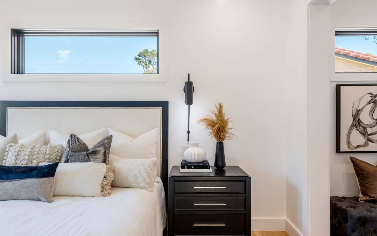 Staged bedroom with a white bed, textured pillows, dark nightstand, and high horizontal windows.