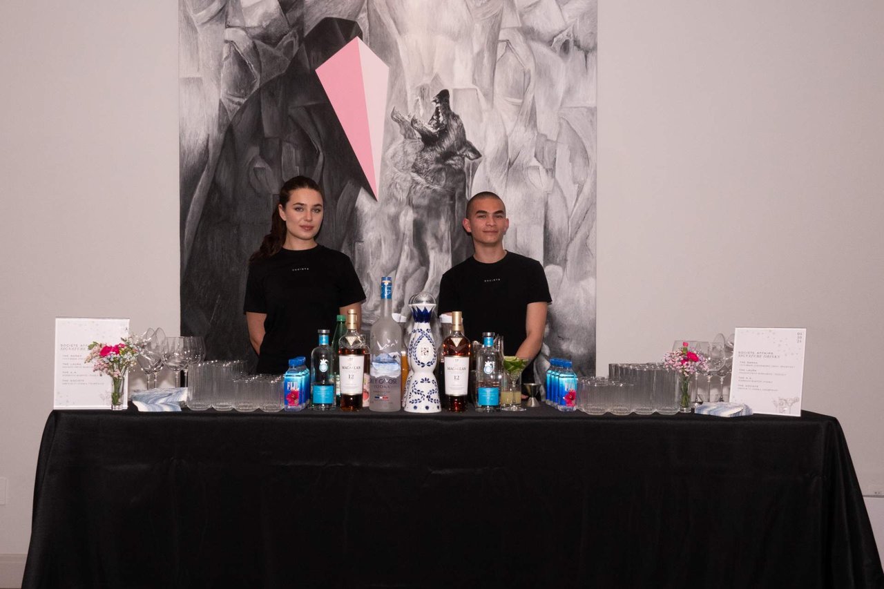 Two people stand behind a drinks table with bottles and glassware in front of a large mural.
